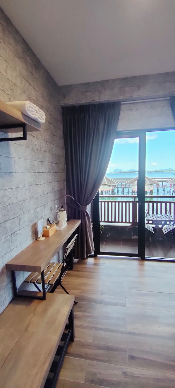 A room with a wooden bench, a small table with a lamp, and a view of a balcony with a railing and outdoor furniture.