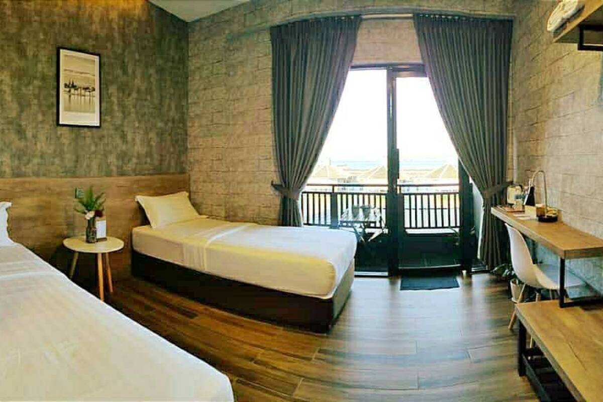 A hotel room with two beds, a small table with a plant, a desk with a chair, and a balcony with a railing.