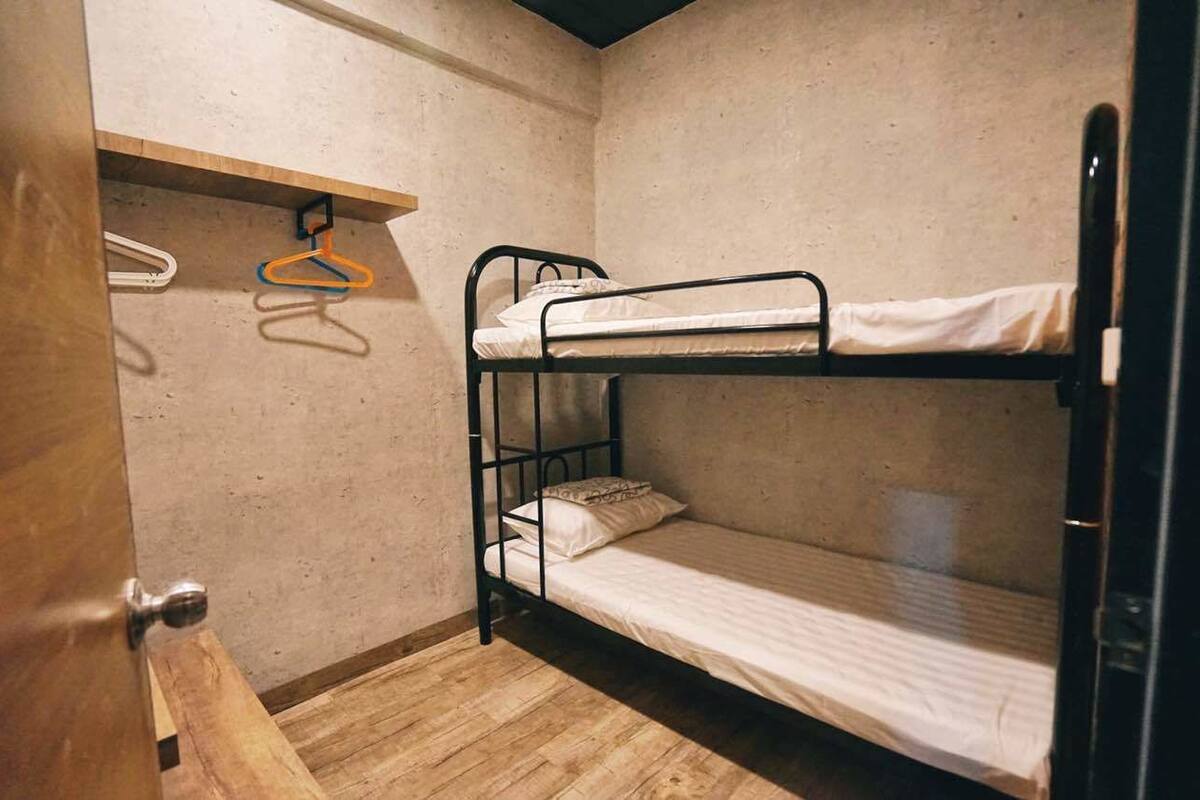 A small, single-bunk room with a wooden floor, a metal bunk bed, and a wooden shelf with hangers.