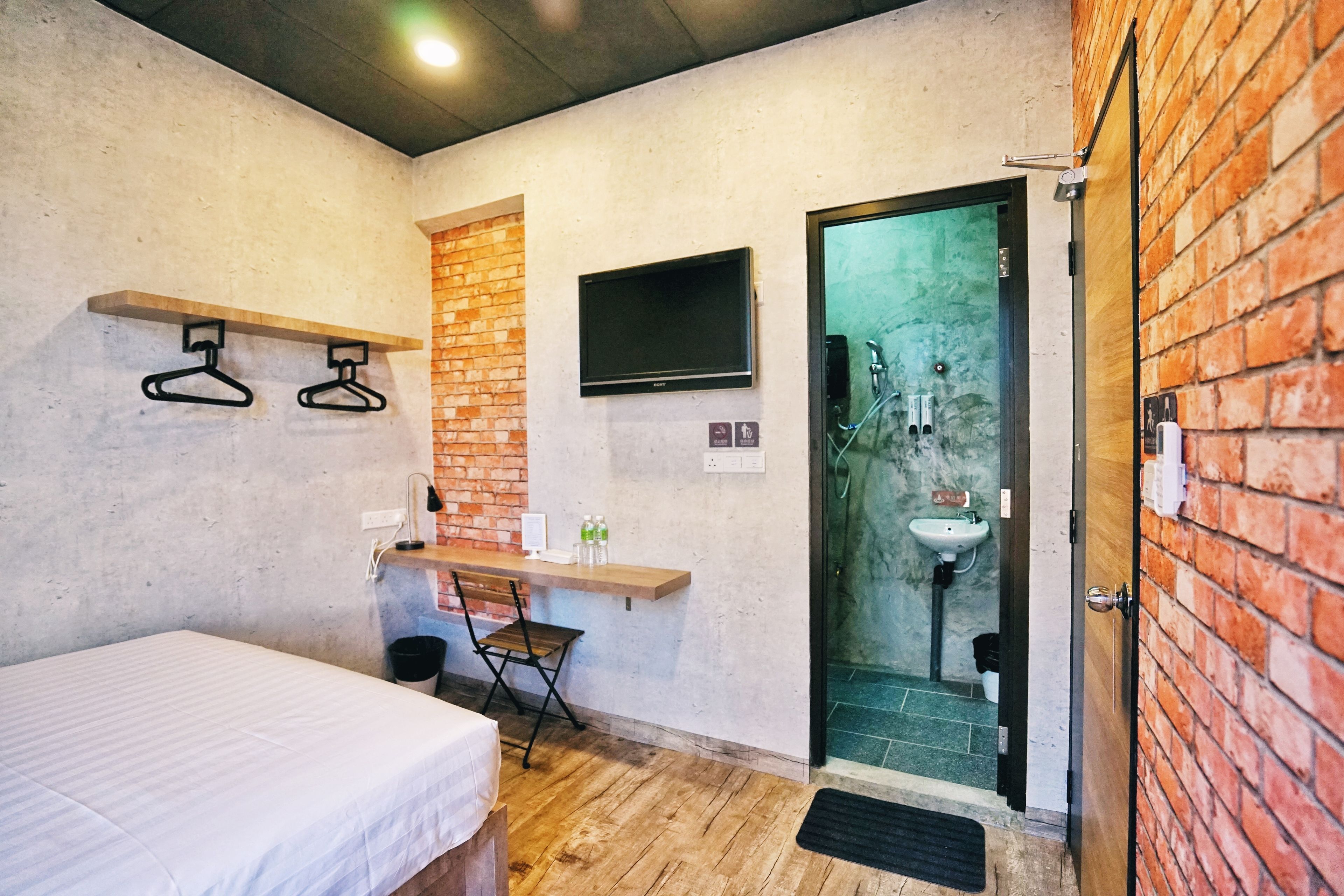 A small, modern room with a bed, a desk, a TV, and a bathroom with a sink and shower.