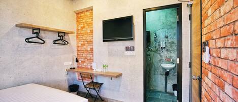 A small, modern room with a bed, a desk, a TV, and a bathroom with a sink and shower.