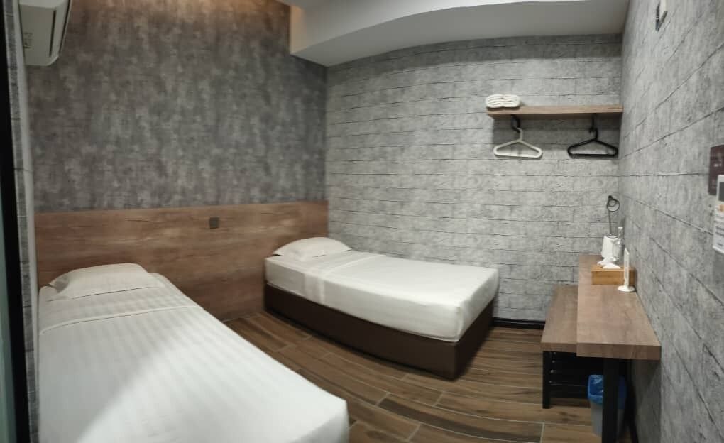 A compact room with two beds, a wooden headboard, a small table with a lamp, and a brick-patterned wall.