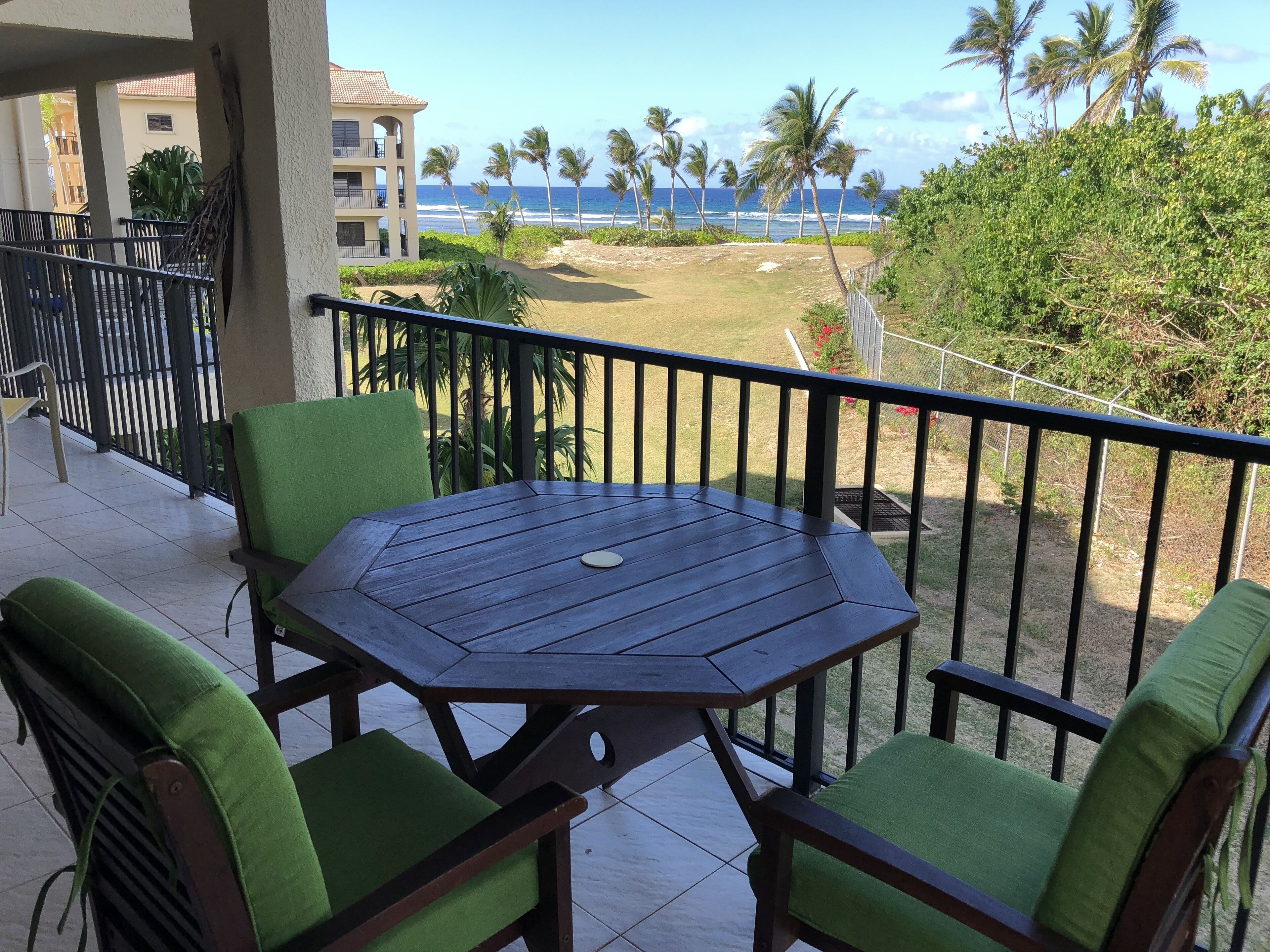A balcony with a wooden table and green cushioned chairs, overlooking a beach with palm trees and a building in the background.