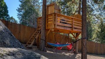 Explore the forest from a rustic playhouse with a ladder leading up to a platform, featuring a hammock between two trees.