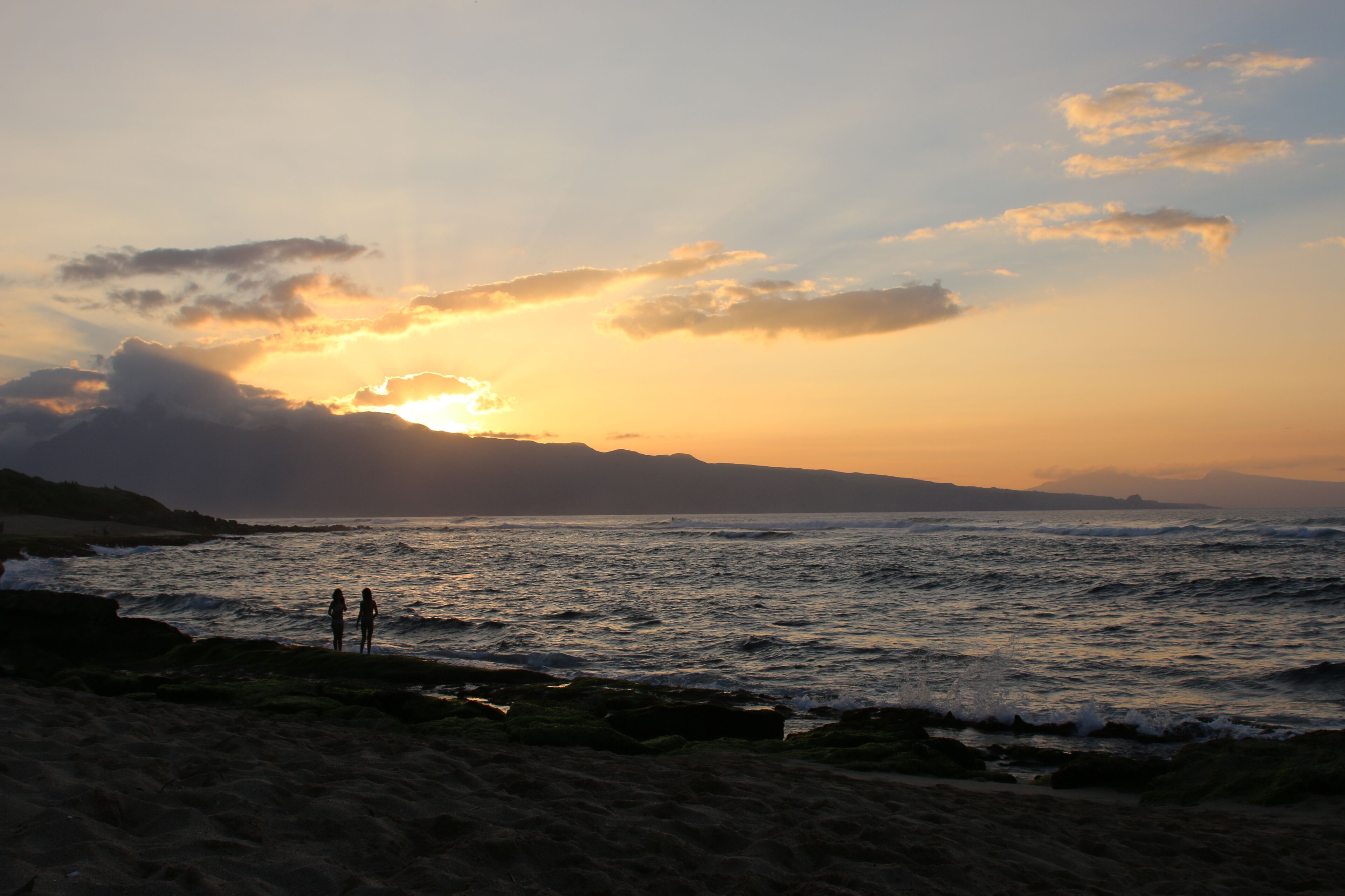 A beach scene with waves, a sunset, and two people standing on the shore.