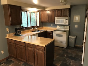 A kitchen with wooden cabinets, a stainless steel sink, a built-in microwave, a stove, and a trash can.