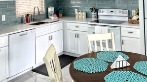 A modern kitchen with white cabinets, a microwave, and a stove. There is a ceiling fan, a window with blinds, and a dining table with blue placemats. - Great rates for September! SIESTA KEY GULF 2 bed 2 bath condo Jamaica Royale 104 (Sarasota)