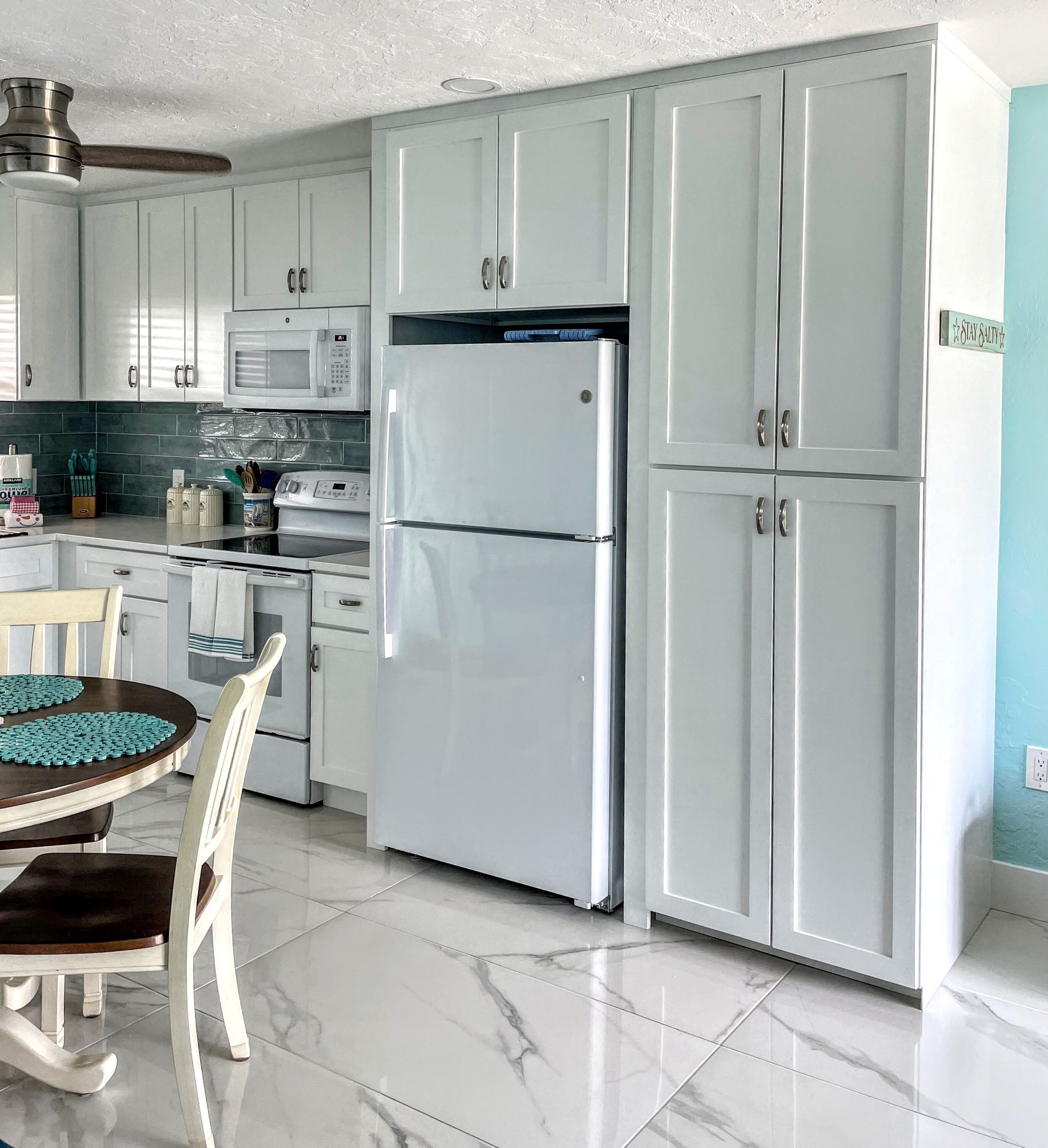 Una cocina con armarios blancos, un refrigerador de doble puerta, un microondas y una mesa de comedor con sillas.