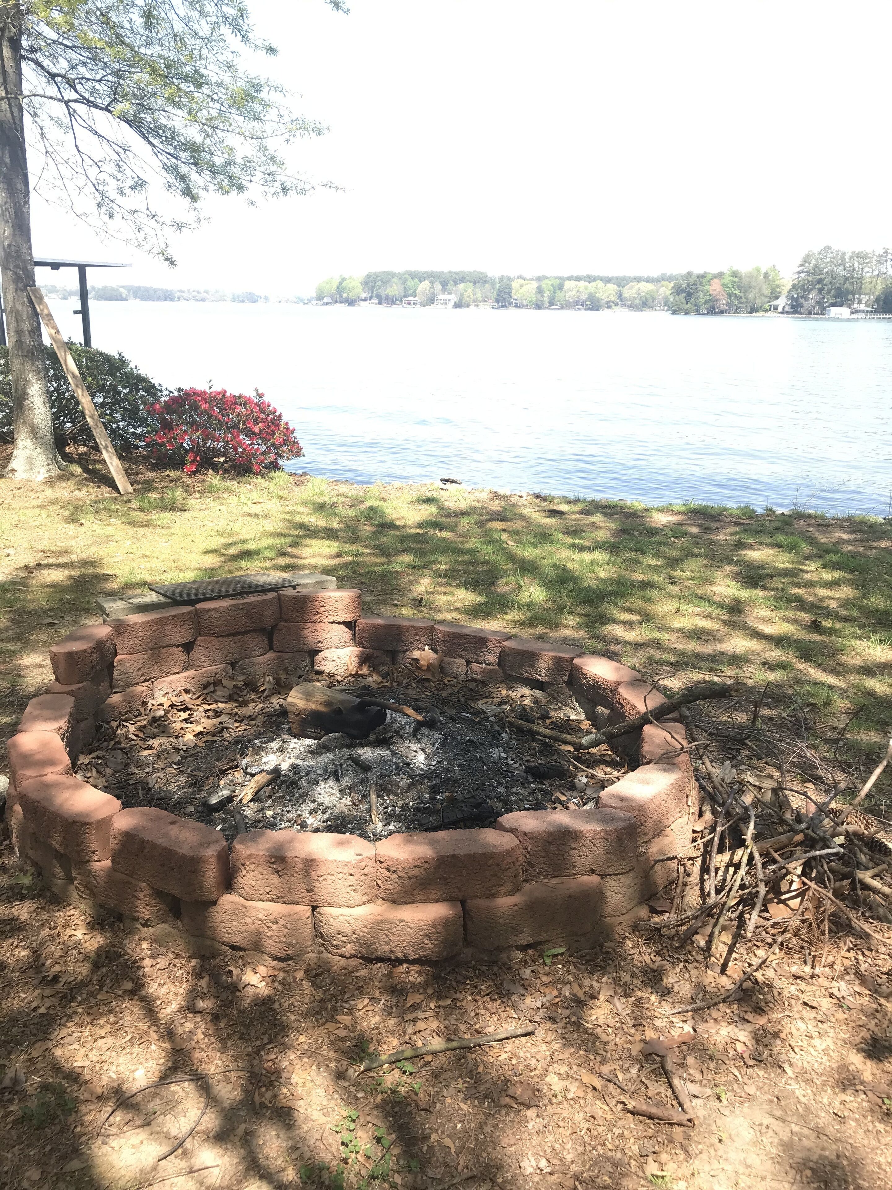 A fire pit area surrounded by a brick border, with a view of a lake and trees in the background.