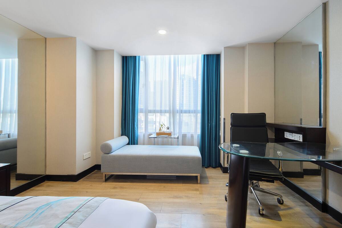 A hotel room with a bed, a desk with a chair, a large window with curtains, and a mirror.