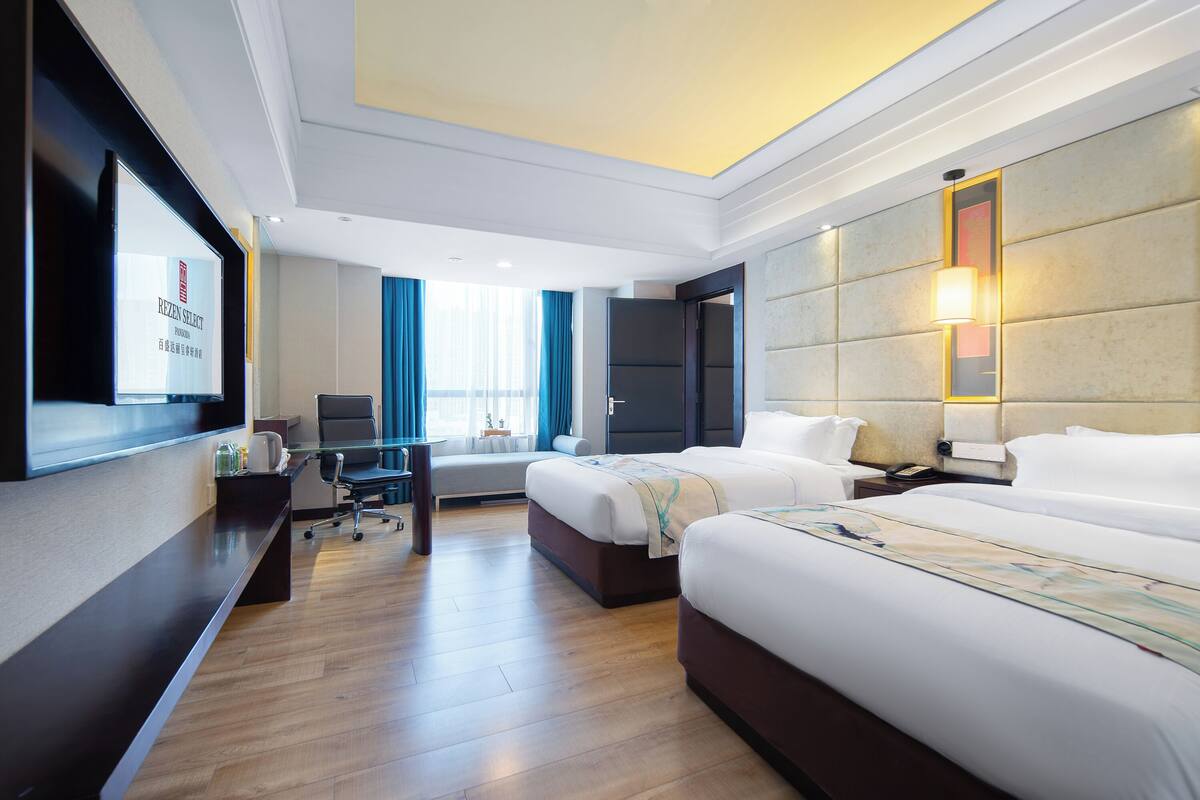 A hotel room with two beds, a desk, a TV, and a large window with curtains.