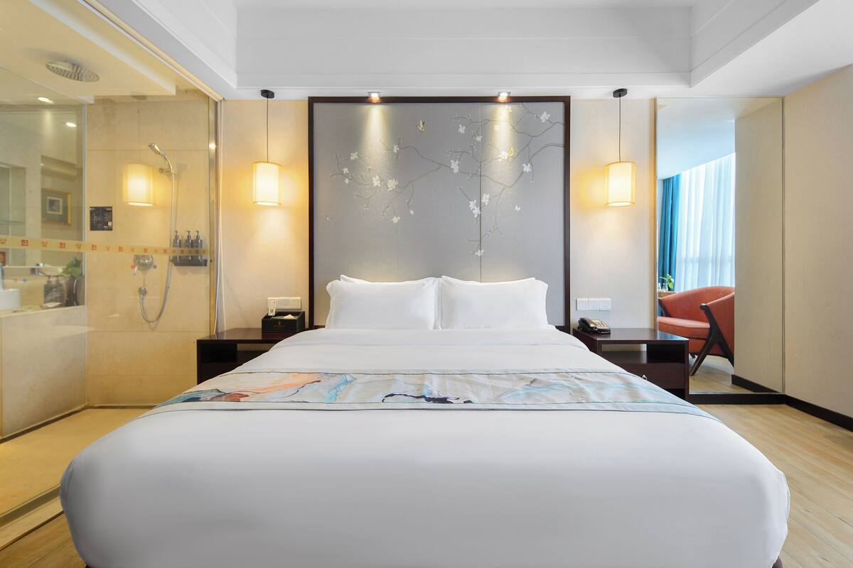 A hotel room with a large bed, a shower area, a desk with a chair, and a decorative wall.