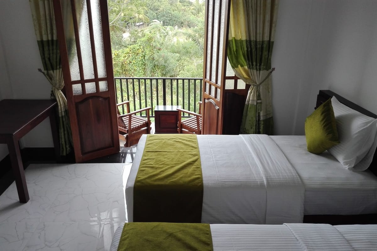 Habitación de hotel con dos camas, un escritorio de madera y un balcón con vistas a zonas verdes.