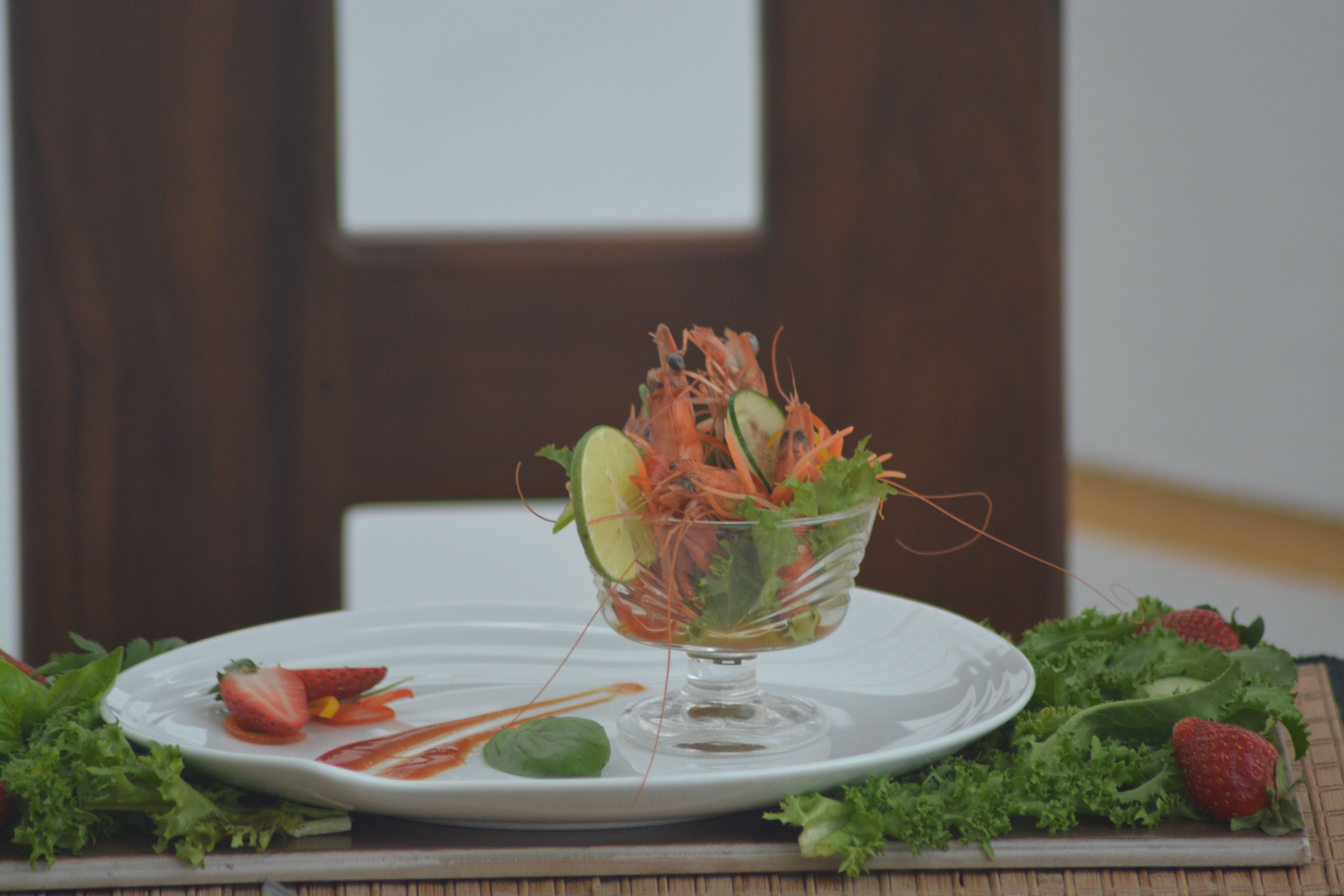 A clear glass bowl filled with a salad on a white plate, surrounded by fresh vegetables and strawberries.