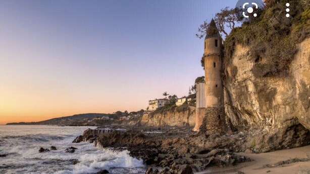 A coastal scene with a rocky beach, waves, and a cliff with a building.