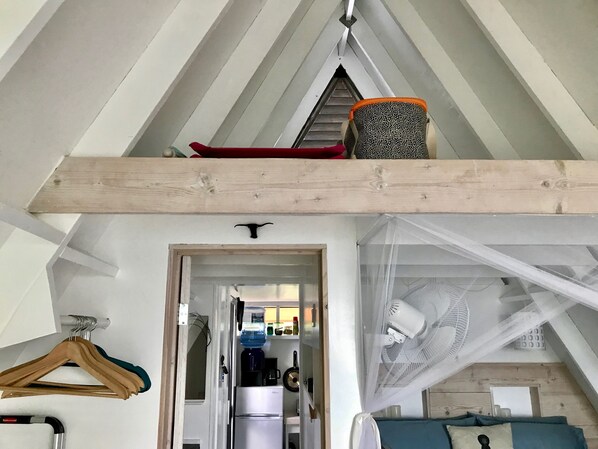 A loft bedroom with a sloped ceiling, a wooden beam ceiling, a bed with a white canopy, a water dispenser, and a kitchenette in the background.
