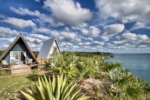 Two A-frame cabins with wooden decks and chairs, surrounded by tropical plants, overlooking a calm sea.