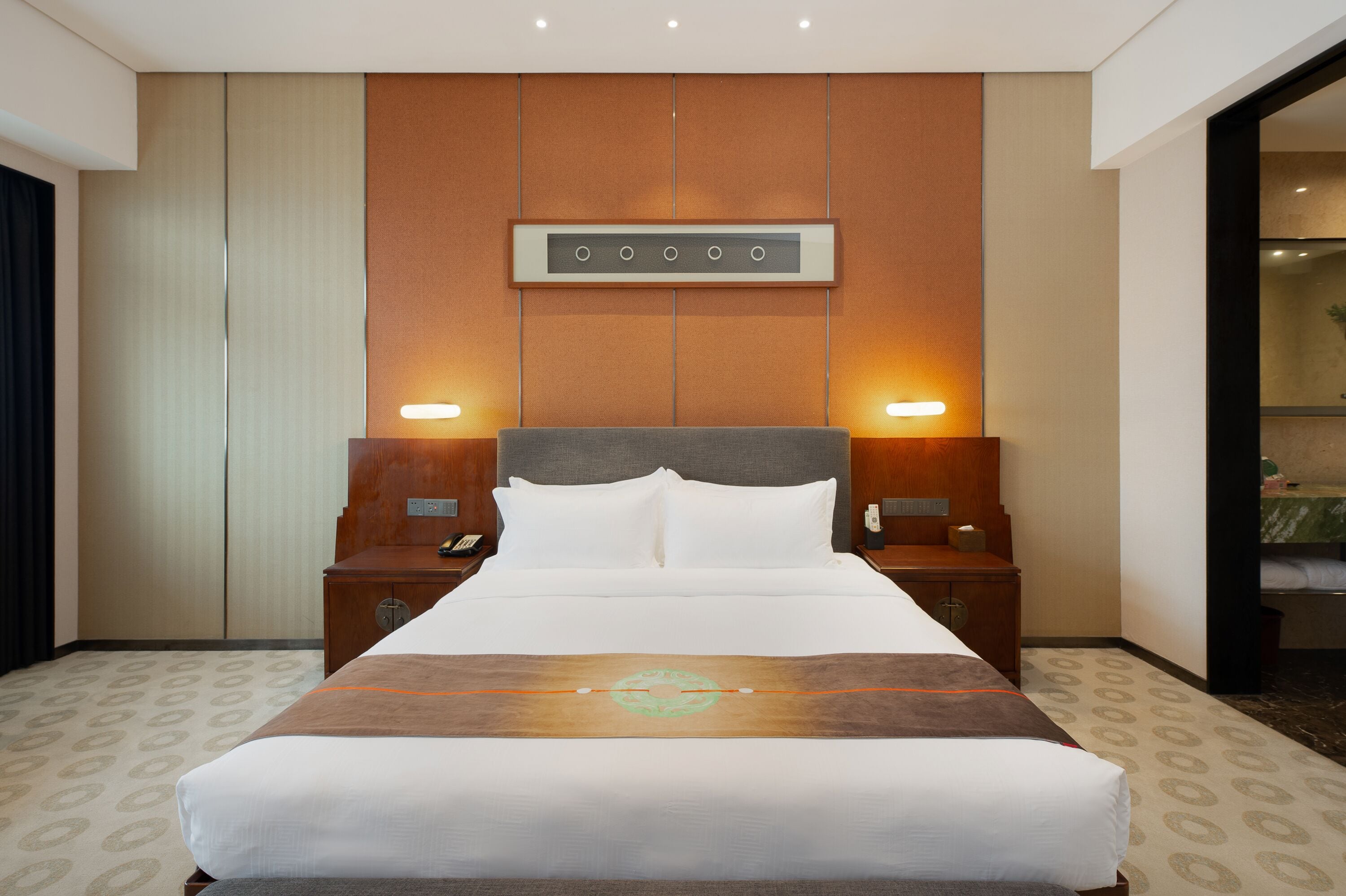 Una habitación de hotel con una cama grande, dos mesitas de noche, un panel de control en la pared y un baño visible a través de una puerta abierta.