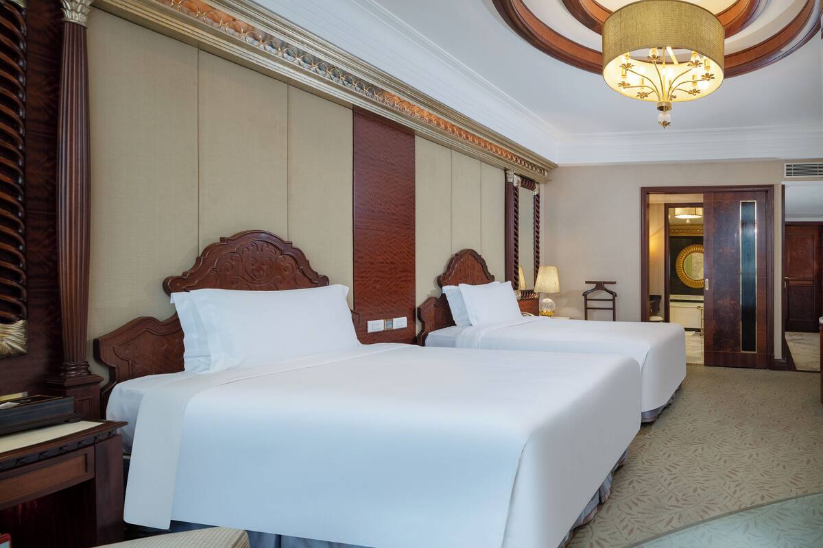A hotel room with two beds, wooden headboards, a chandelier, and a carpeted floor.