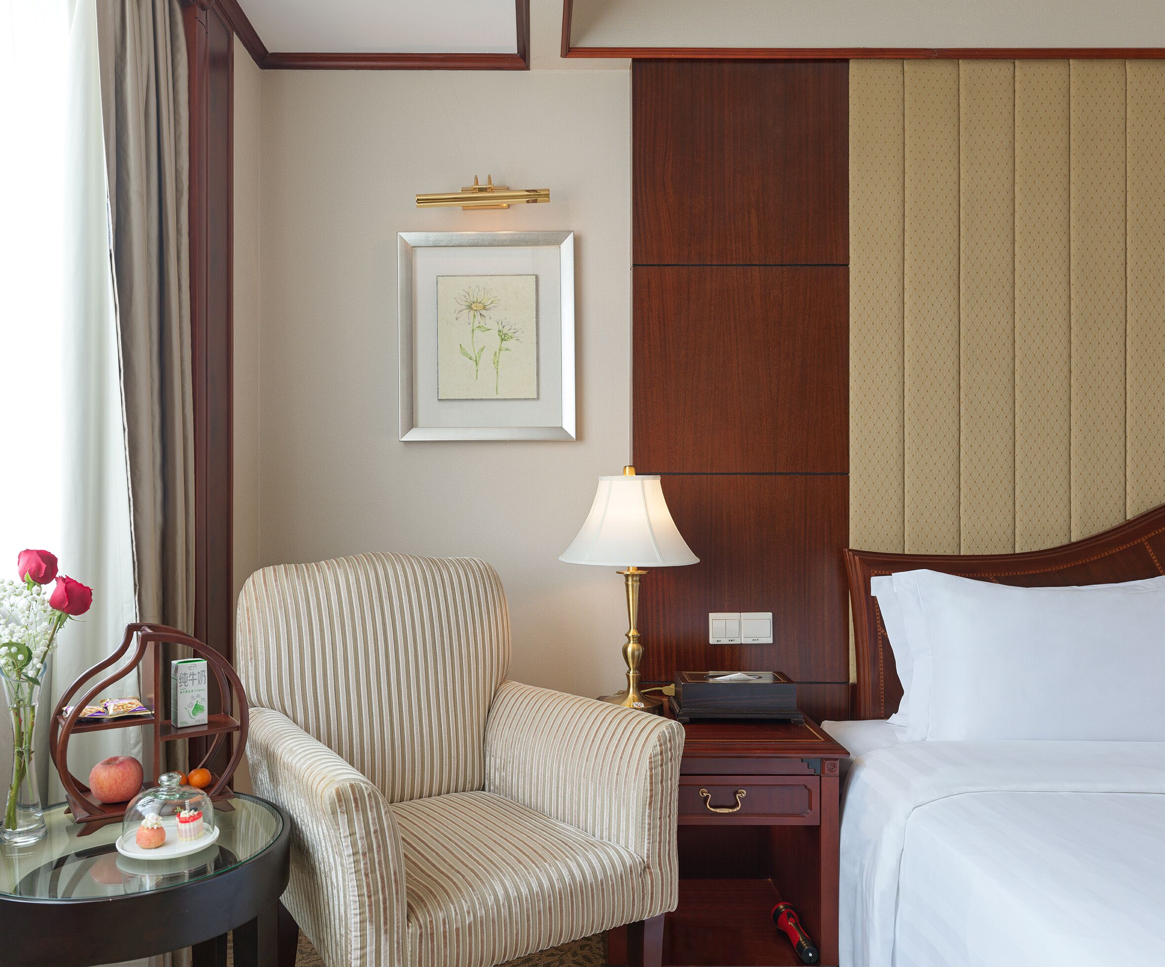 A hotel room with a bed, a bedside table with a lamp, a chair, a small table with a vase of flowers, and a framed picture on the wall.