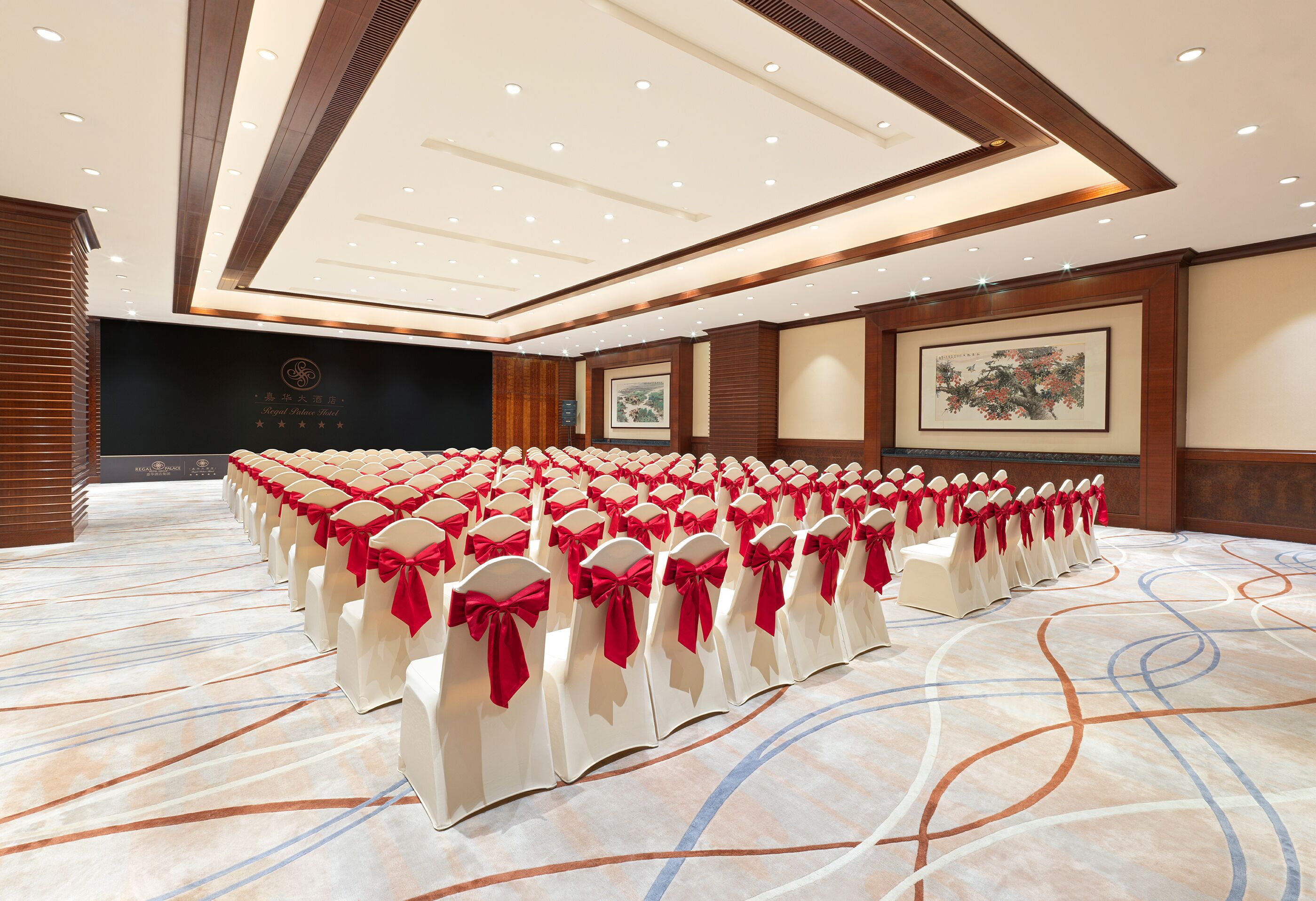 Une salle de conférence avec des rangées de chaises blanches ornées de rubans rouges, une moquette à motifs et des panneaux de bois sur les murs.