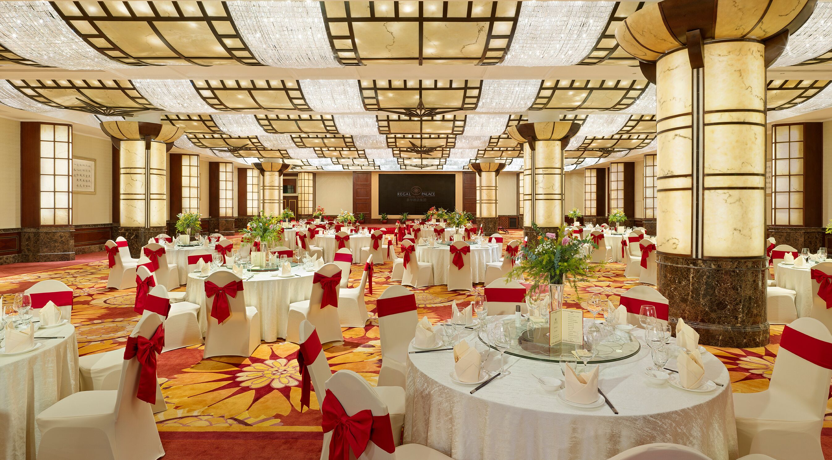 A banquet hall with round tables, white chairs with red sashes, and a chandelier ceiling.