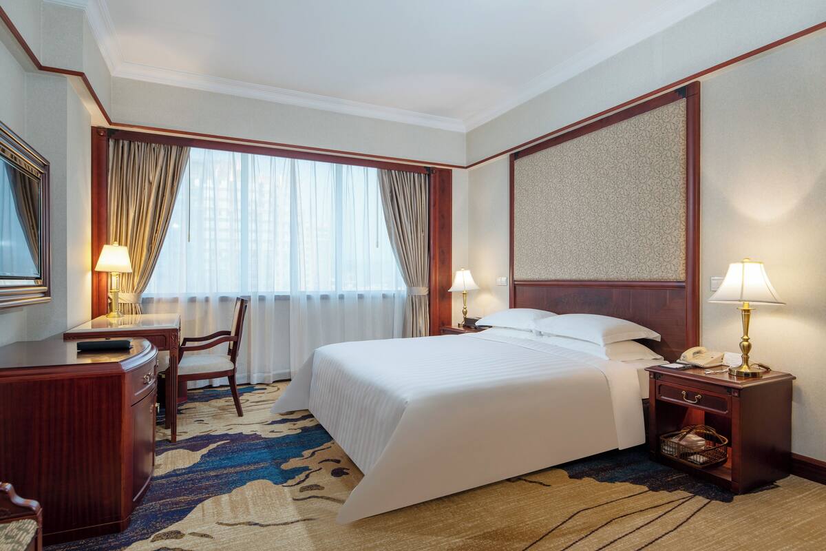 A hotel room with a large bed, a desk, a chair, two bedside lamps, and a patterned rug.