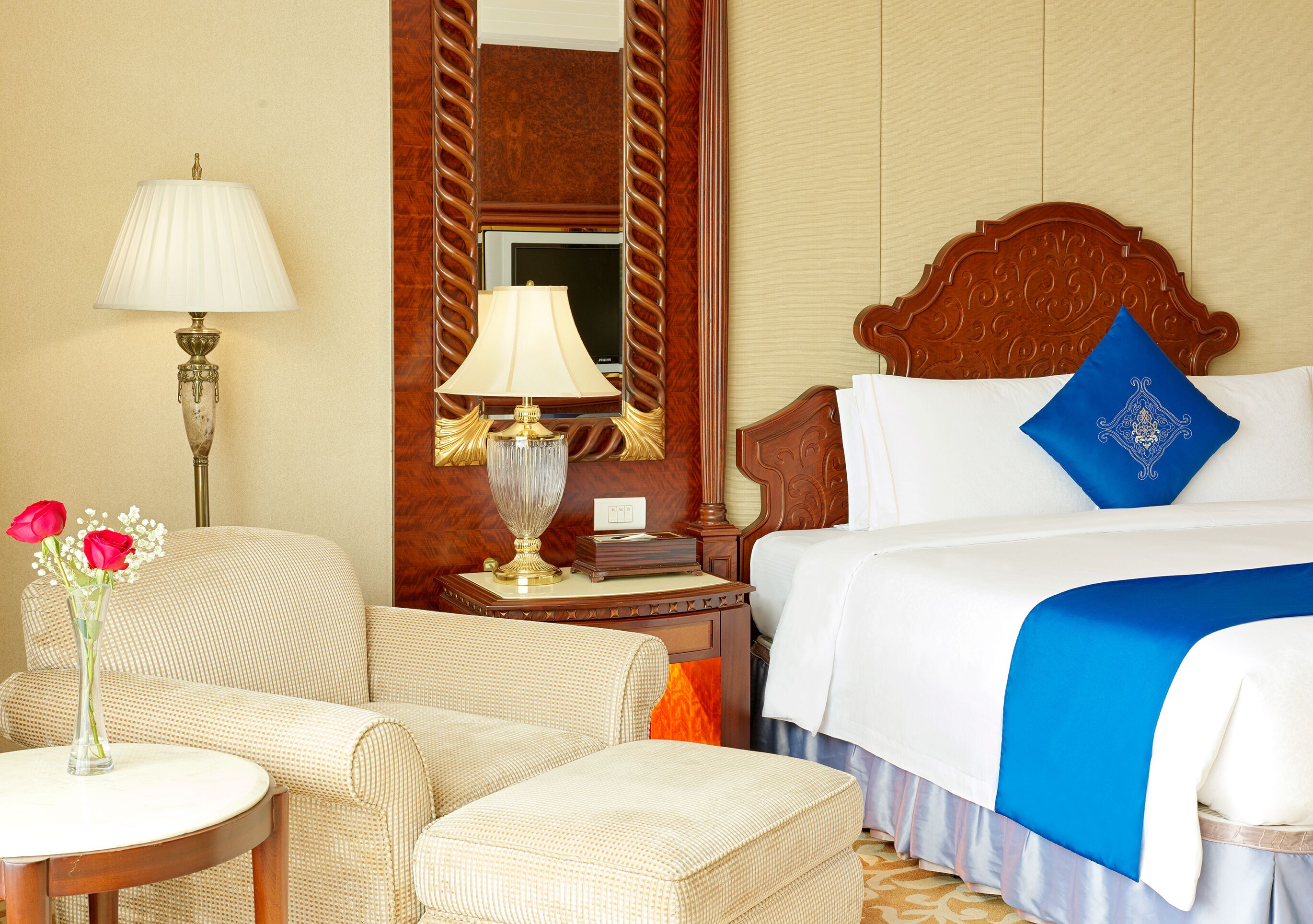 Una habitación de hotel con cama, dos sillas, una mesita de noche con lámparas, un espejo y un jarrón con flores.