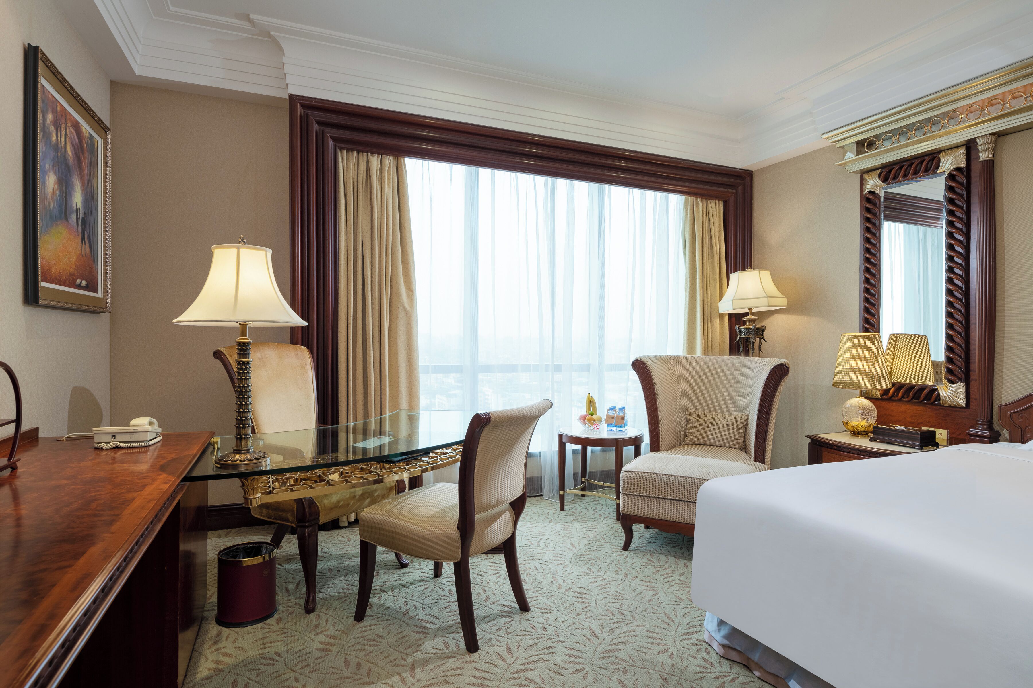 A hotel room with a large bed, two armchairs, a desk with a lamp, a small table with a lamp, a painting on the wall, and a large window with curtains.