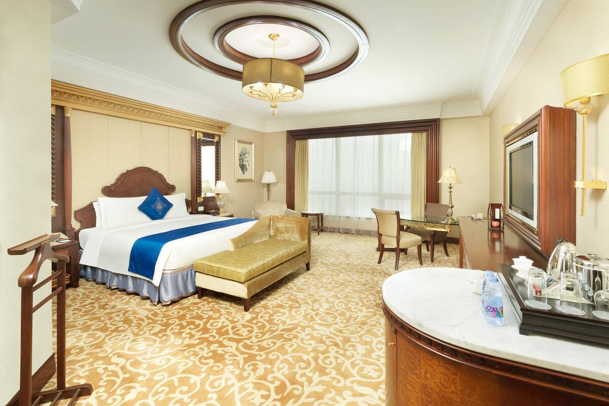 A hotel room with a large bed, a sitting area, a desk, a television, and a bathroom with a marble countertop.