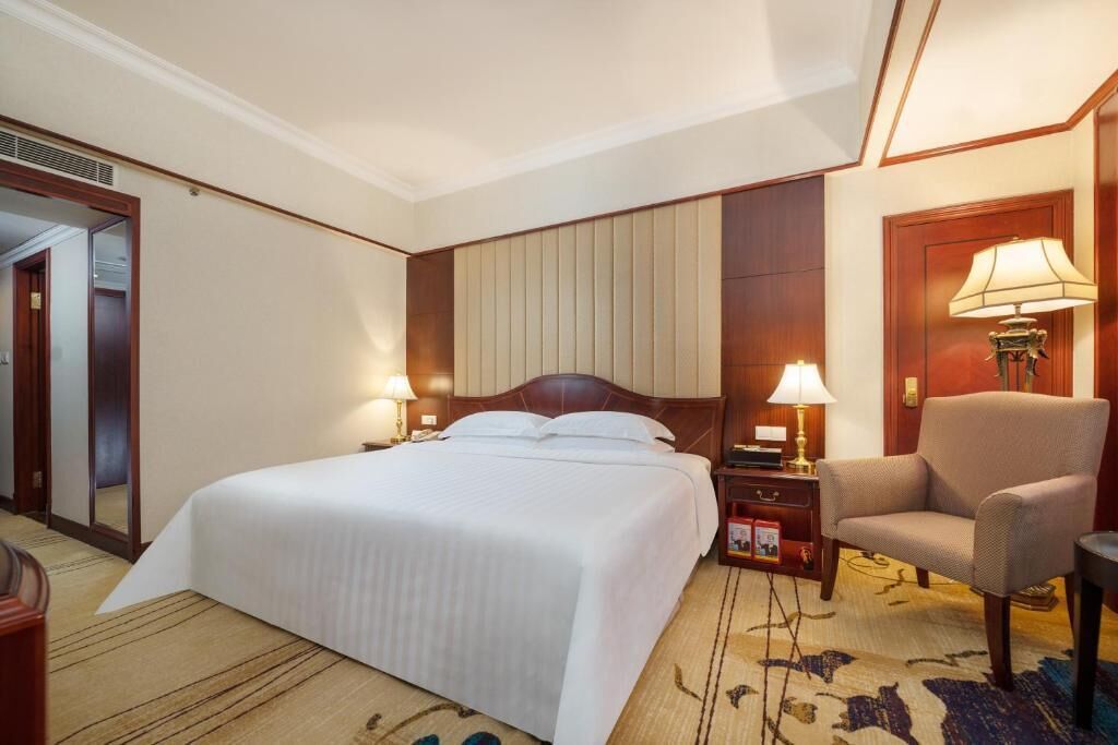 Una habitación de hotel con una cama grande, dos mesitas de noche con lámparas, una silla y piso alfombrado.