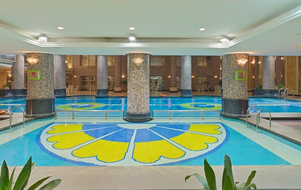 Una piscina interior con diseño de suelo de baldosas, rodeada de zonas de estar y columnas con motivos de mosaico.