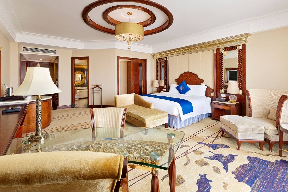 A hotel room with a large bed, a sitting area with a sofa and armchairs, a glass coffee table, and a chandelier.