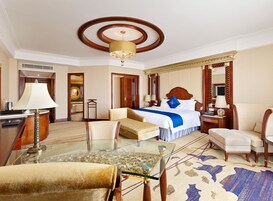 Una habitación de hotel con una cama grande, una zona de estar con sofá y sillones, una mesa de centro de vidrio y un candelabro.