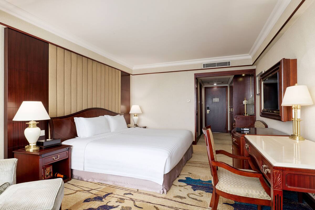 A hotel room with a large bed, two bedside tables with lamps, a desk with a chair, a mirror, and a carpeted floor.