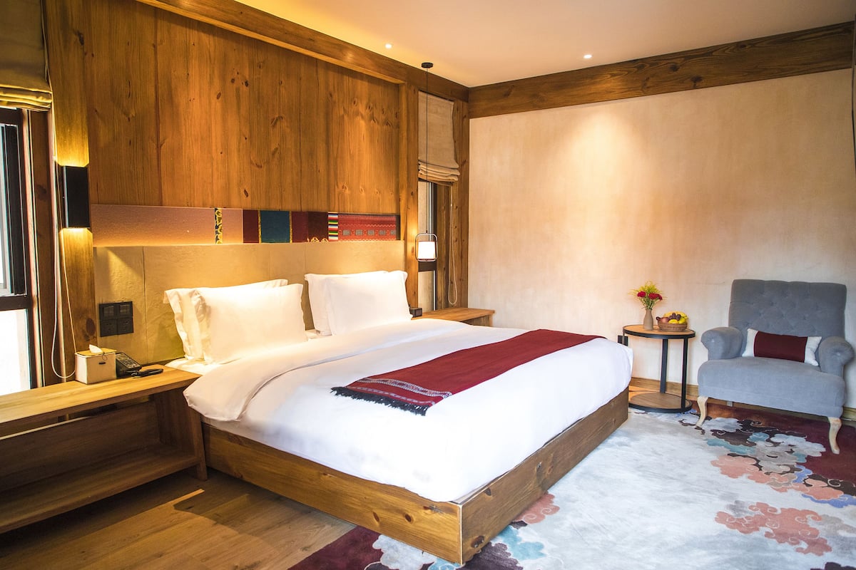 Une chambre d’hôtel avec une tête de lit en bois, un grand lit avec du linge de lit blanc, un plaid rouge, un fauteuil gris et une petite table ronde sur laquelle est posé un vase de fleurs.