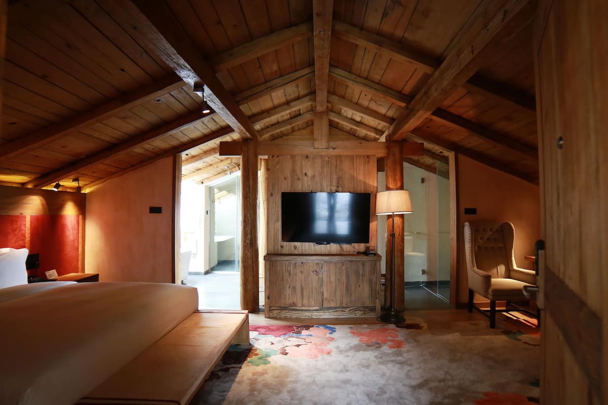 Une chambre confortable située sous les combles, avec un plafond en bois, un grand lit, un téléviseur fixé au mur et un fauteuil.