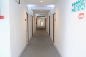 A long, narrow hallway with doors on both sides, a fire exit sign, and a fire alarm on the wall.