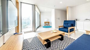 A modern living room with a large window, a blue sofa, a wooden coffee table, and a small side table. - Blanc Bleu Pension (Seogwipo)