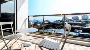 A balcony with two white chairs and a small round table, overlooking a coastal area with a bridge, boats, and buildings. - Blanc Bleu Pension (Seogwipo)