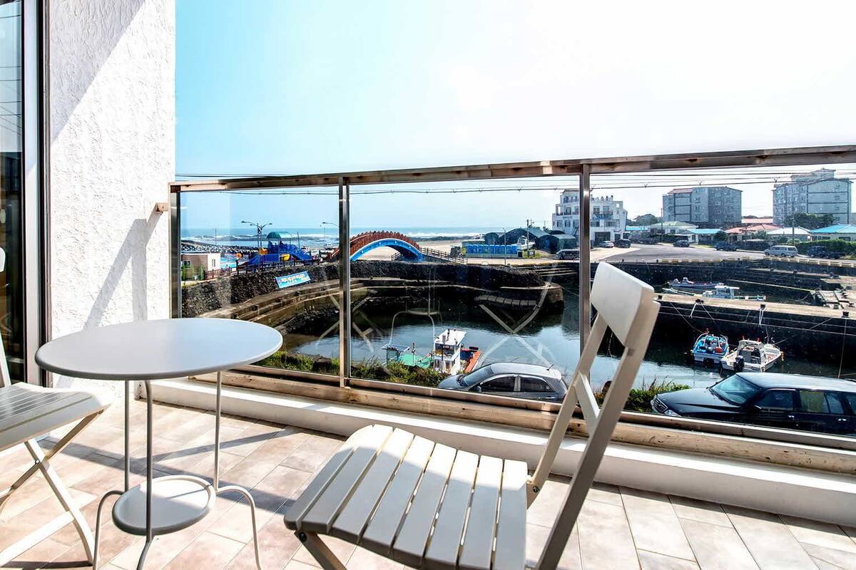 A balcony with two white chairs and a small round table, overlooking a coastal area with a bridge, boats, and buildings.