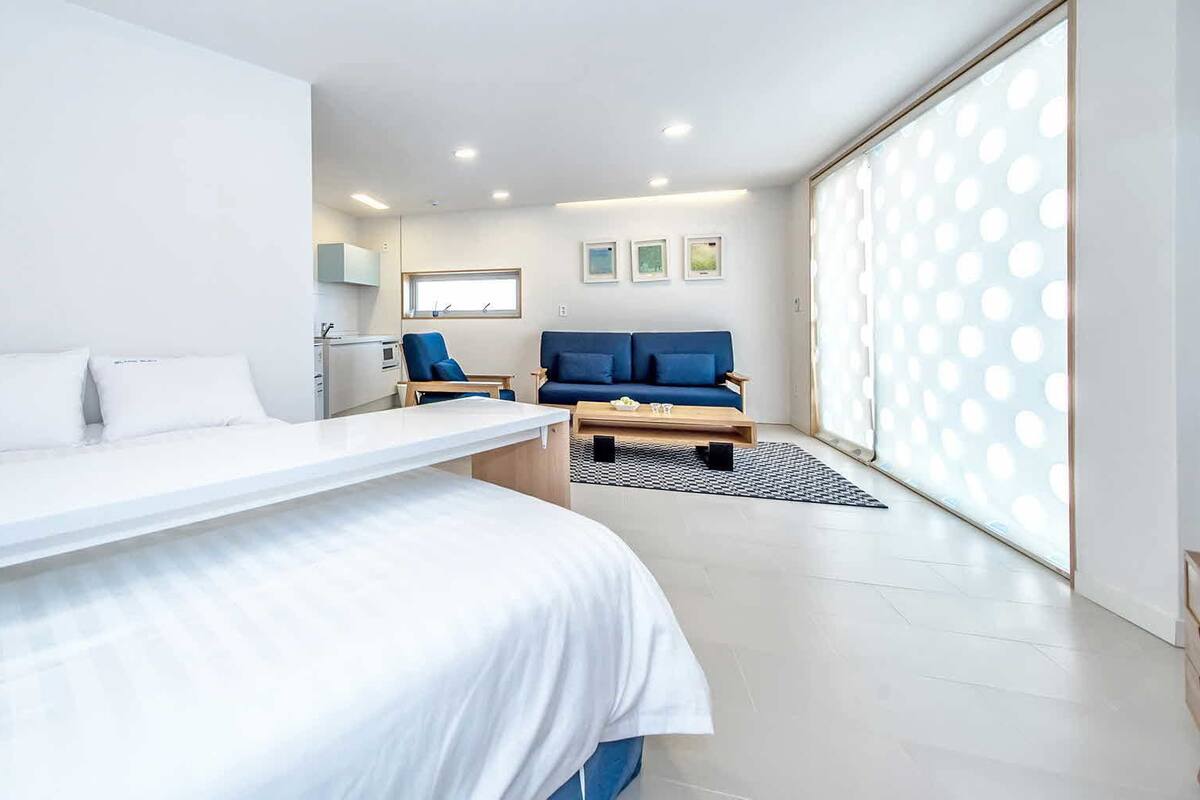 A modern hotel room with a large bed, a desk, a blue sofa, and a view of the city through large windows.