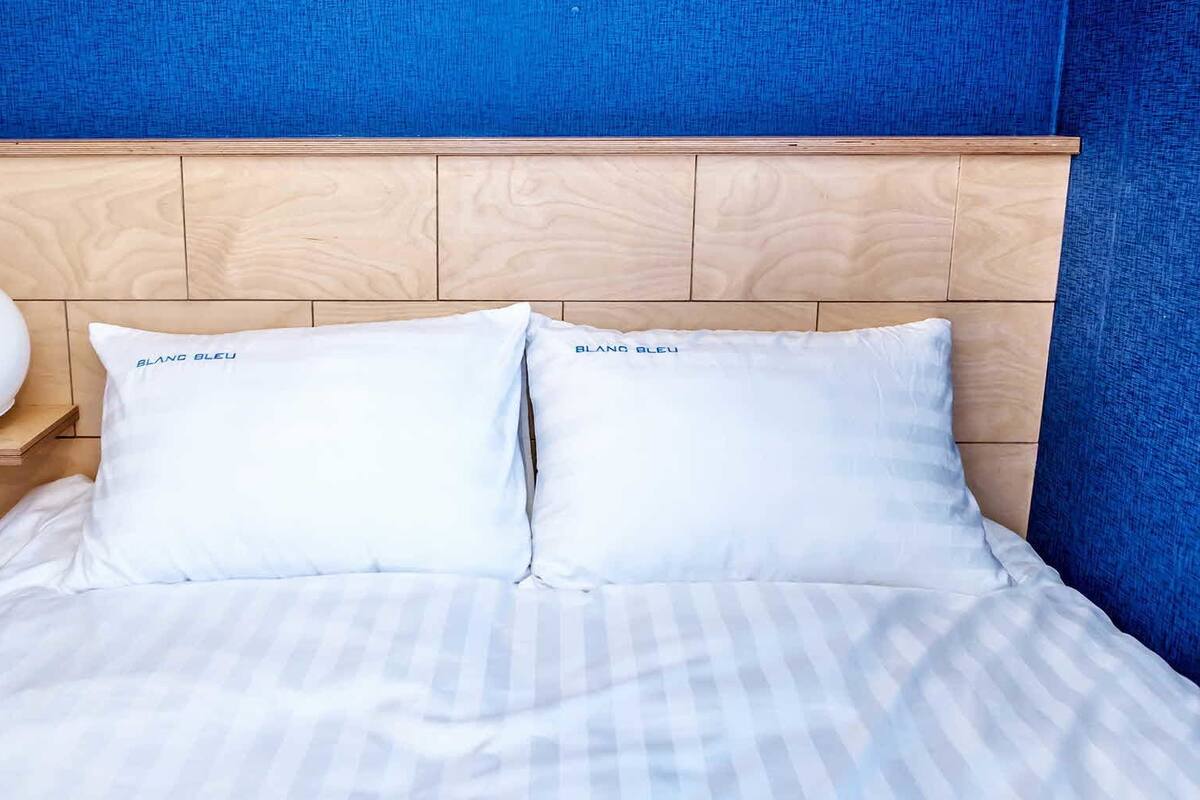 A bed with white bedding and two pillows labeled "BLANG BLEU" against a wooden headboard.