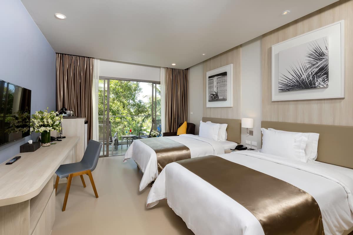 Quarto de hotel com duas camas, uma mesa de trabalho com cadeira, televisão e varanda com vista para o verde.