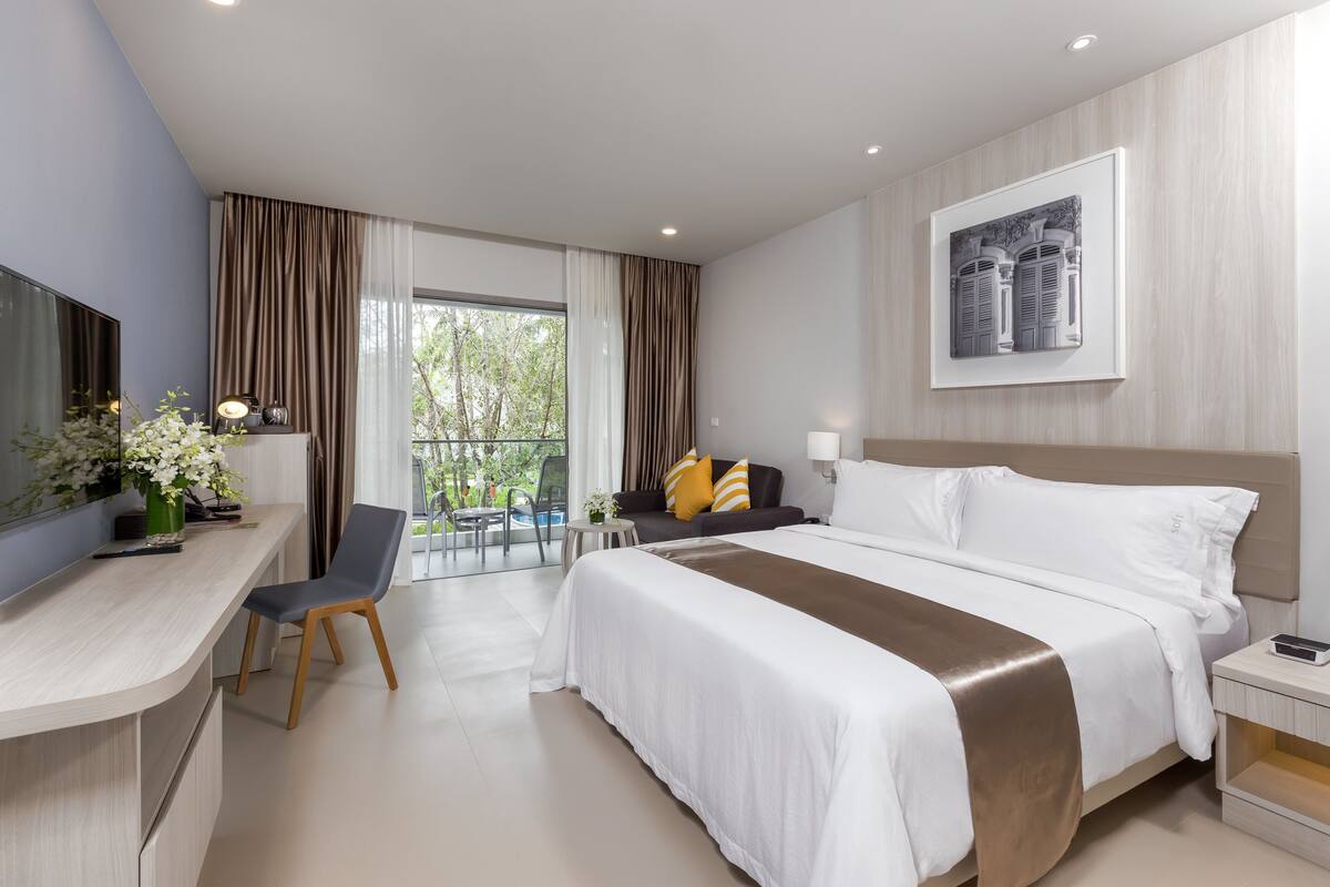 Quarto de hotel moderno com uma cama grande, uma escrivaninha com cadeira, uma mesinha e uma varanda com vista para o verde.