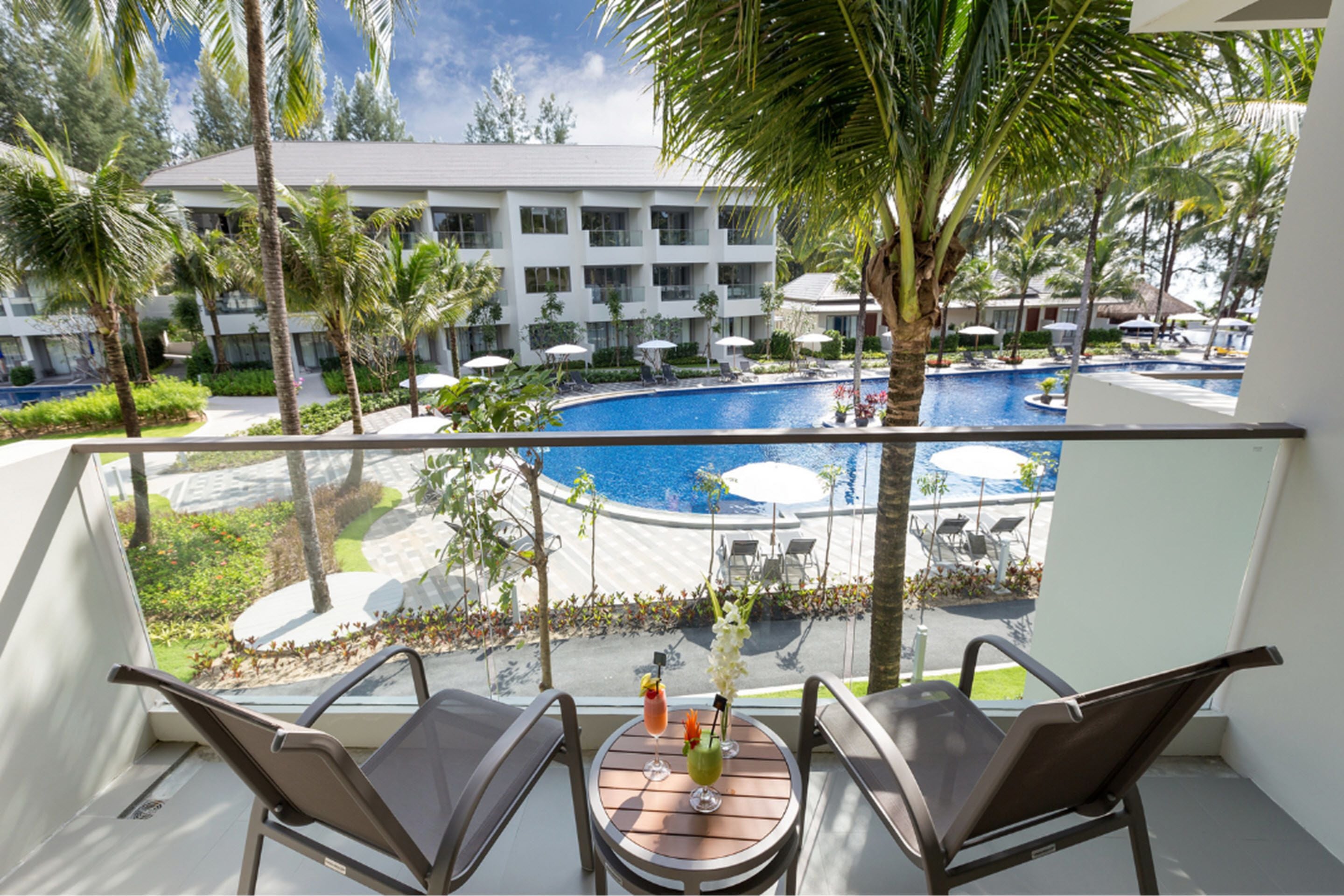 Un balcón con dos sillas y una mesa, con vistas a una zona de piscina con sombrillas y palmeras.