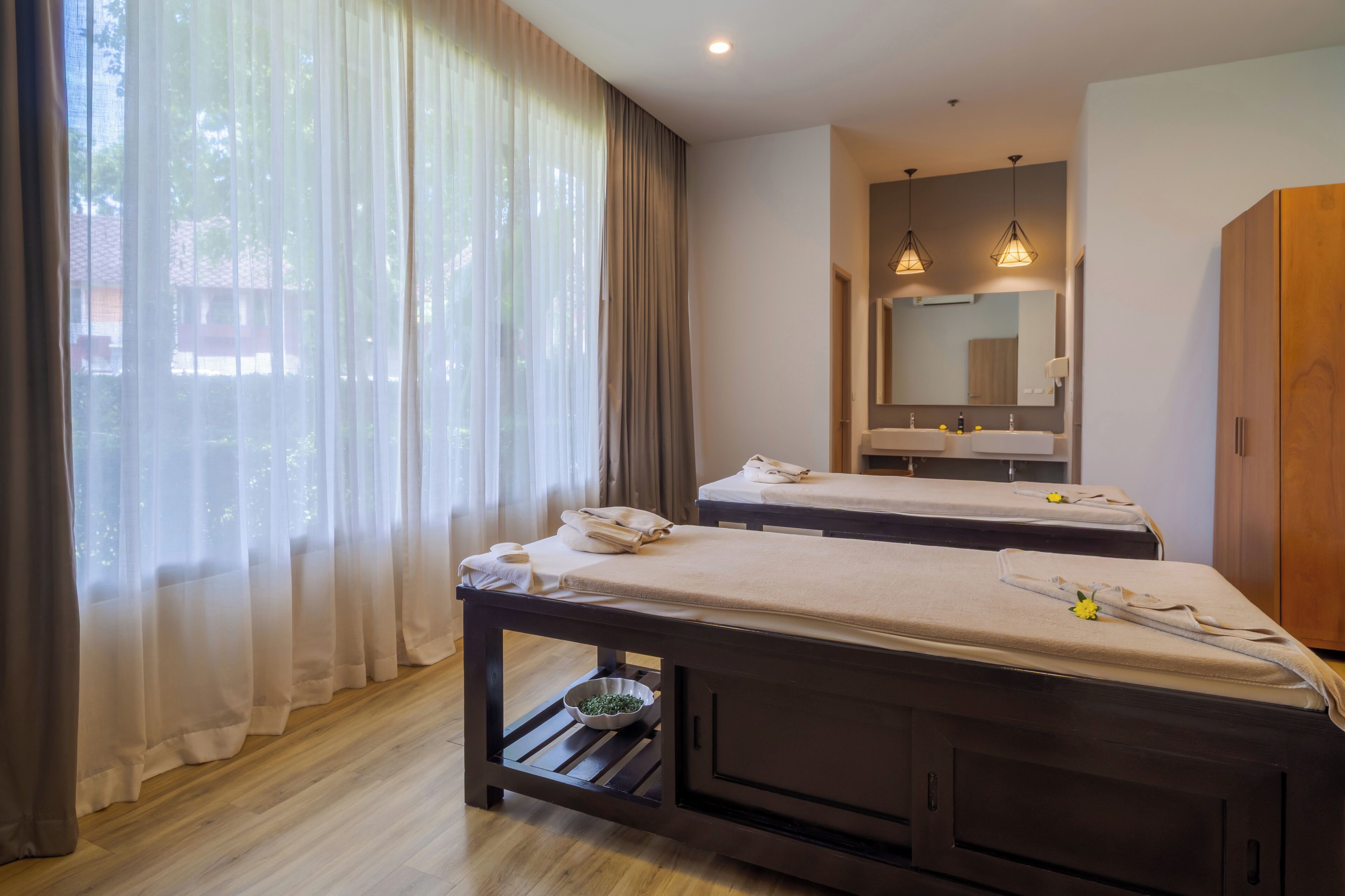 Una habitación con dos camas, un armario de madera, un espejo y una ventana con cortinas traslúcidas.