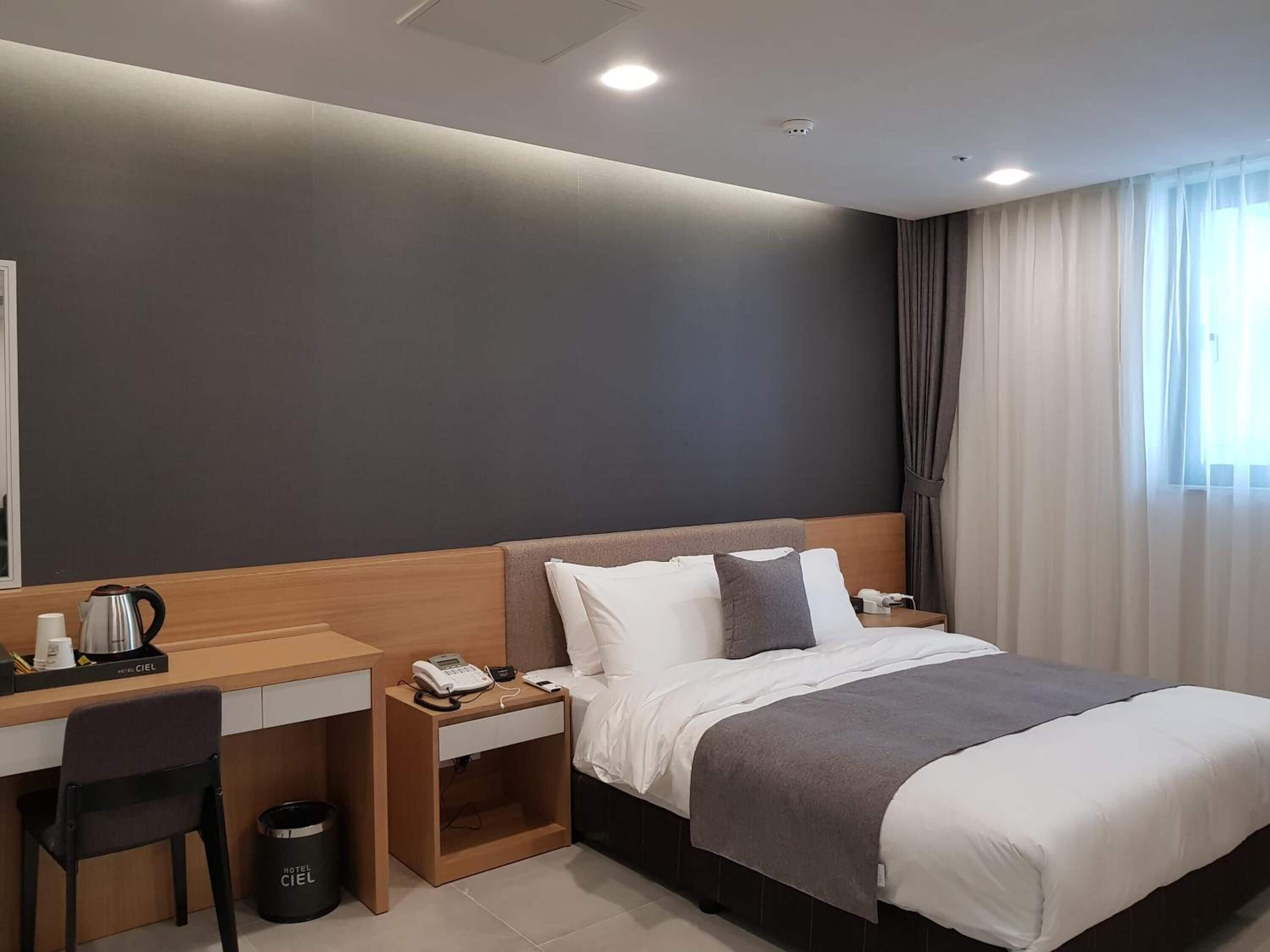 Een moderne hotelkamer met een bed, een bureau en een stoel. Er staat een telefoon op het bureau en een prullenbak naast de stoel.
