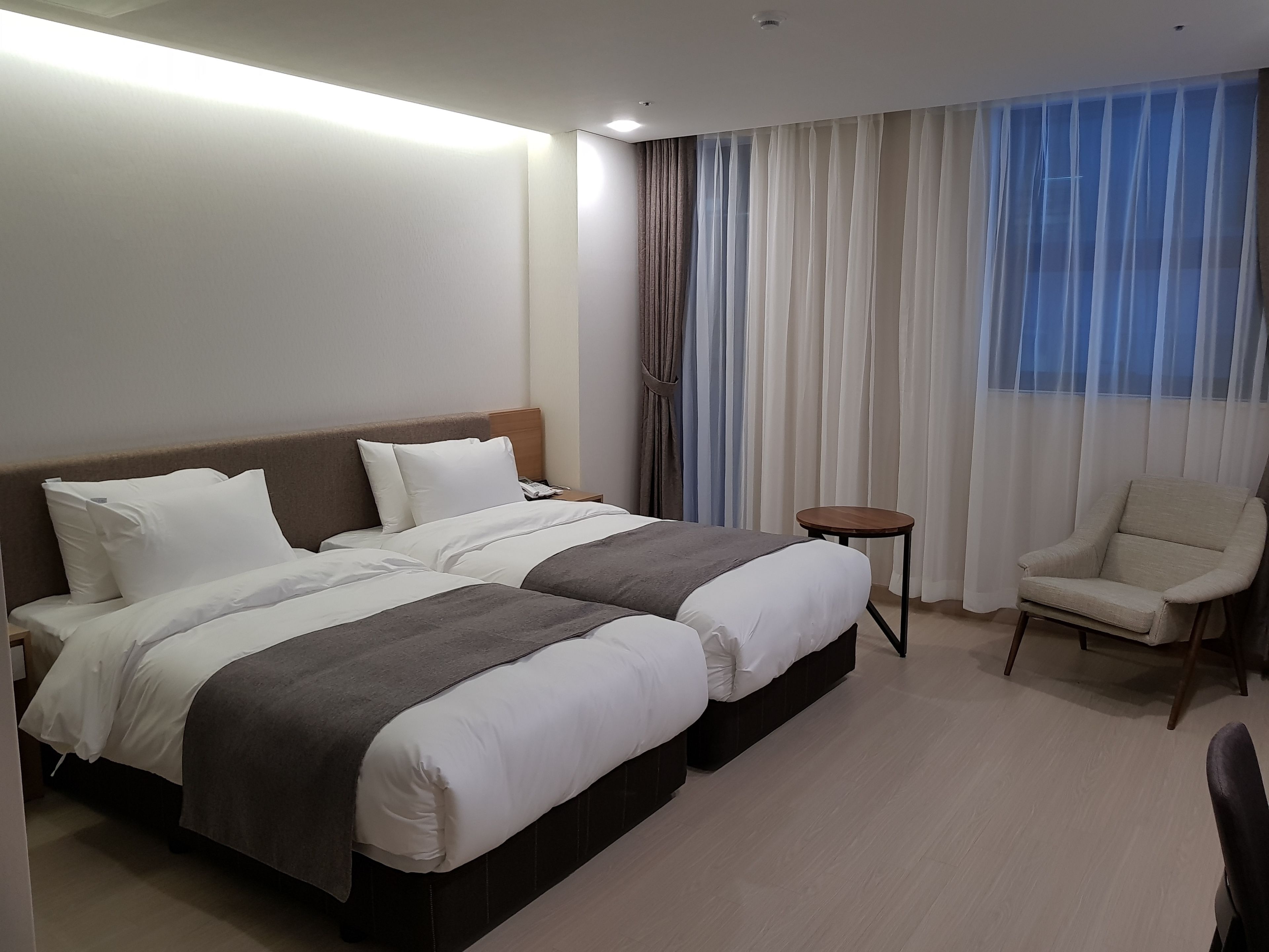Habitació d'hotel amb un llit doble, una taula petita rodona, una cadira i un gran finestral amb cortines.
