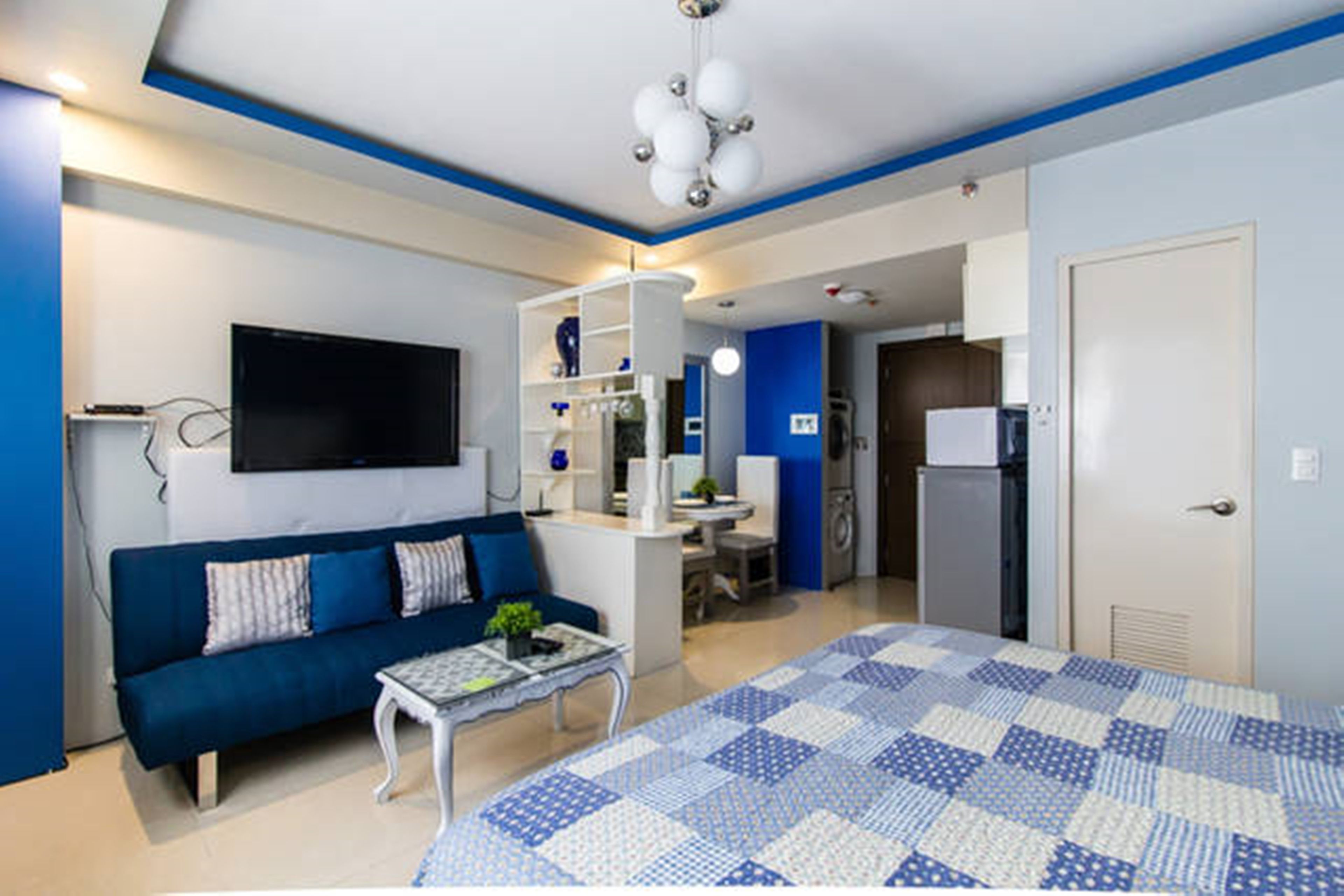 Una sala moderna con un sofá azul, una mesa de centro blanca y una cama grande con colcha azul y blanca. Hay un televisor de pantalla plana montado en la pared, una pequeña mesa con una planta y una cocina visible al fondo.